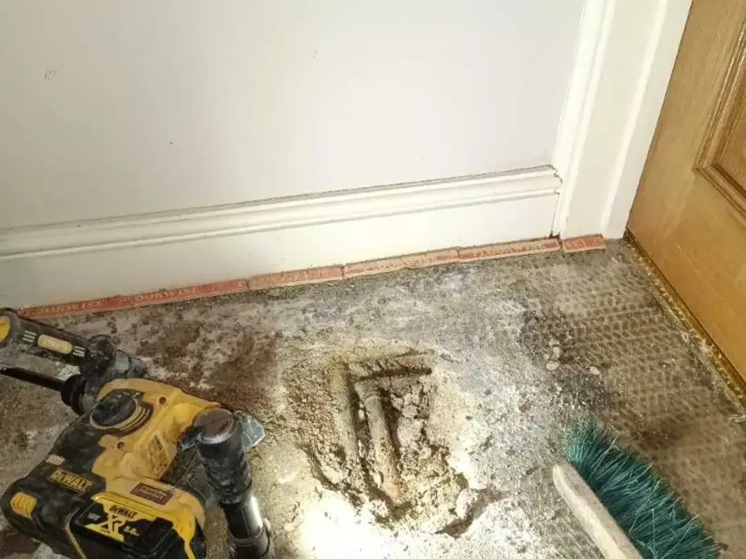 A hole broken out of a hallway floor beside the skirting, with a breaker drill next to it